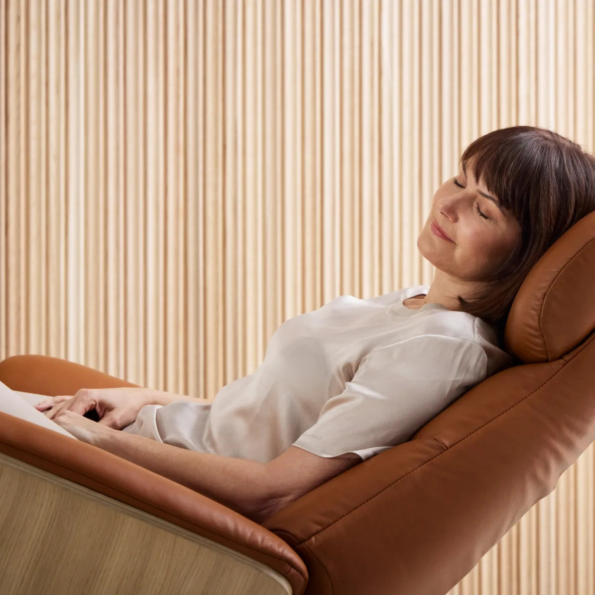 A woman is reclining comfortably in a Stressless Adam upholstered in Paloma New Cognac leather