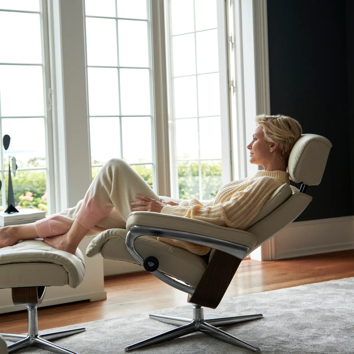 Une femme est confortablement installée dans un fauteuil Stressless Erik monté sur un piètement Cross, près d'une grande fenêtre