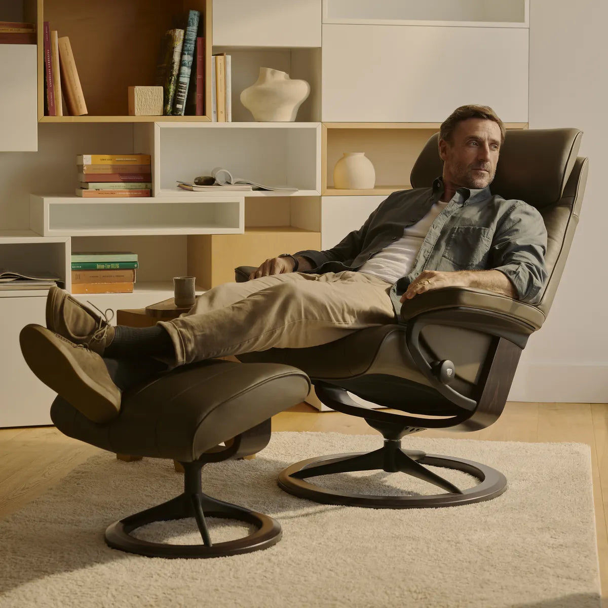 A man relaxes in the reclining position in a Stressless Paul with a footstool, upholstered in Velaro Forest Green leather on a Signature base