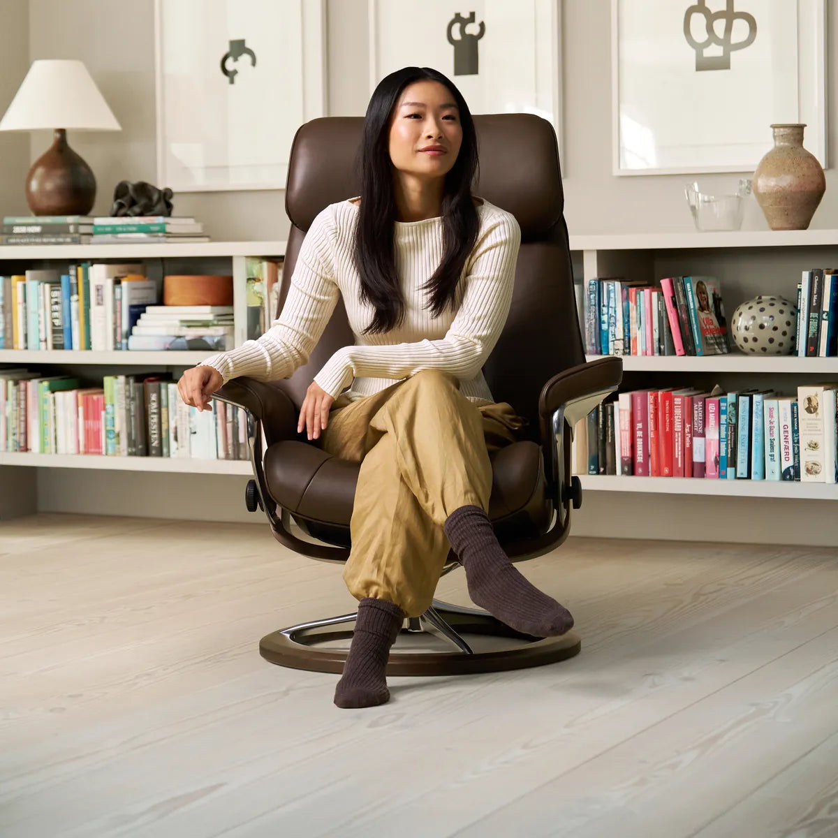 A woman is sitting comfortably in a Stressless Paul upholstered in Paloma Chestnut leather on a Signature base with walnut, in front of a bookcase