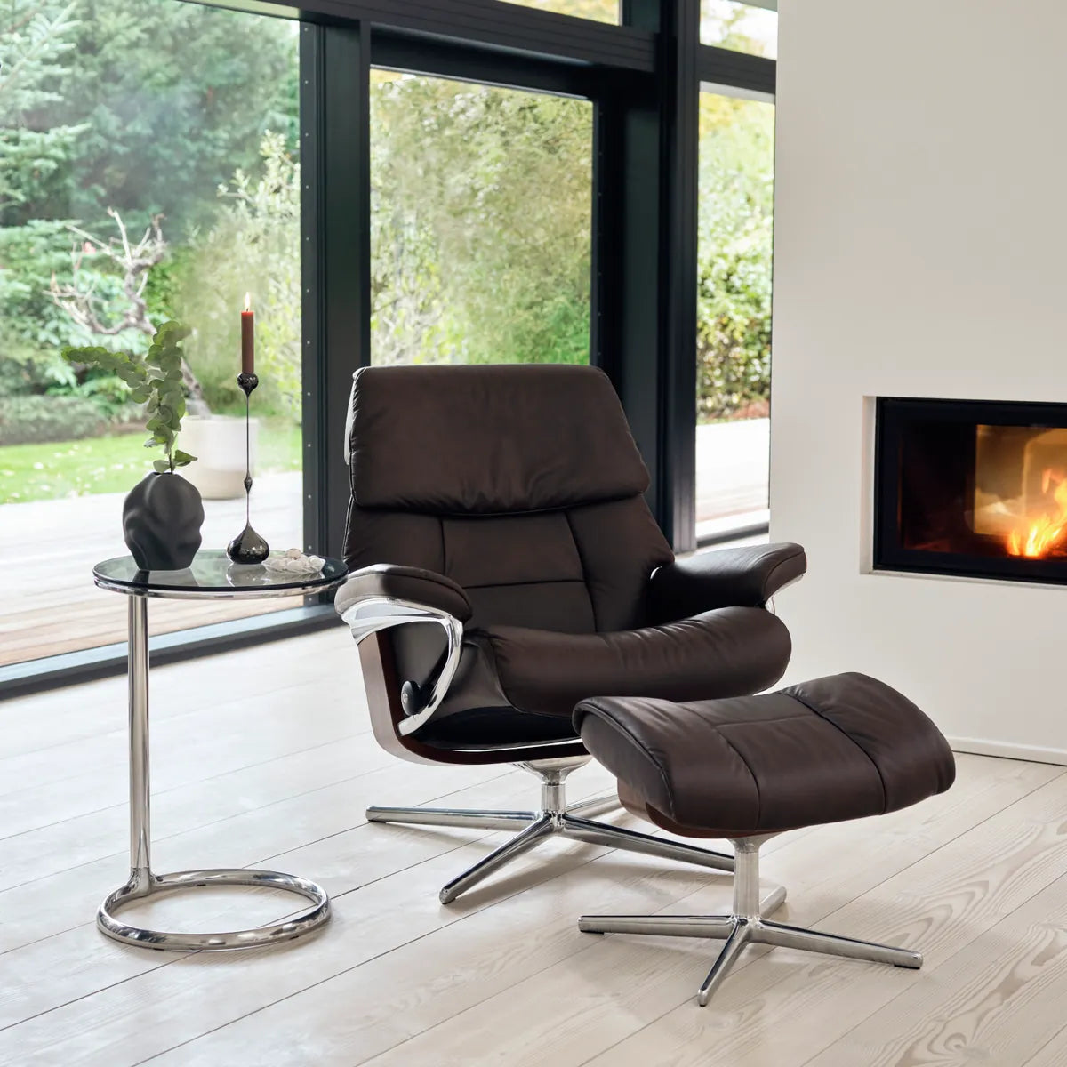 Stressless Ruby met voetenbank in Paloma Chocolate leder op een Cross onderstel, naast een open haard en groot raam