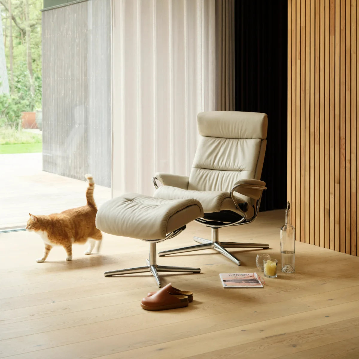 Stressless Tokyo met verstelbare hoofdsteun in Paloma Fog leder met voetenbank in een lichte kamer met houten wand, met een kat ernaast
